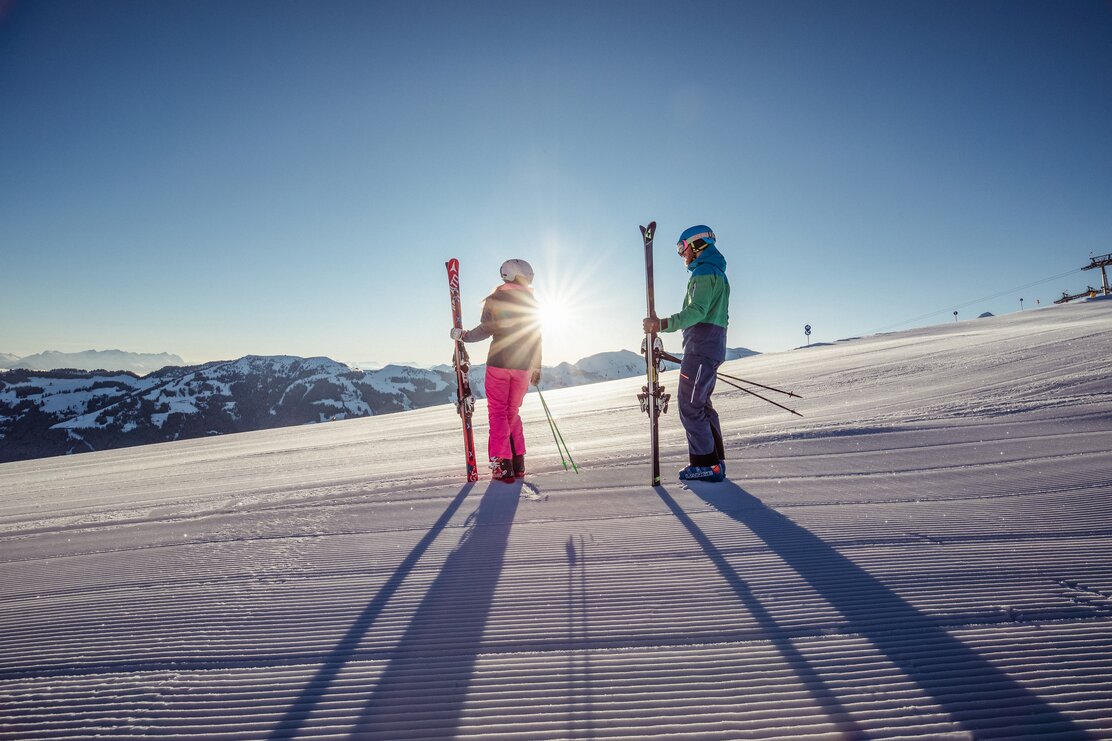 Eine Frau und ein Mann stehen samt Skiausrüstung auf der frisch präparierten Piste, die Sonne scheint und der Schnee glitzert   | © Ski Juwel Alpbachtal Tourismus | shootandstyle 