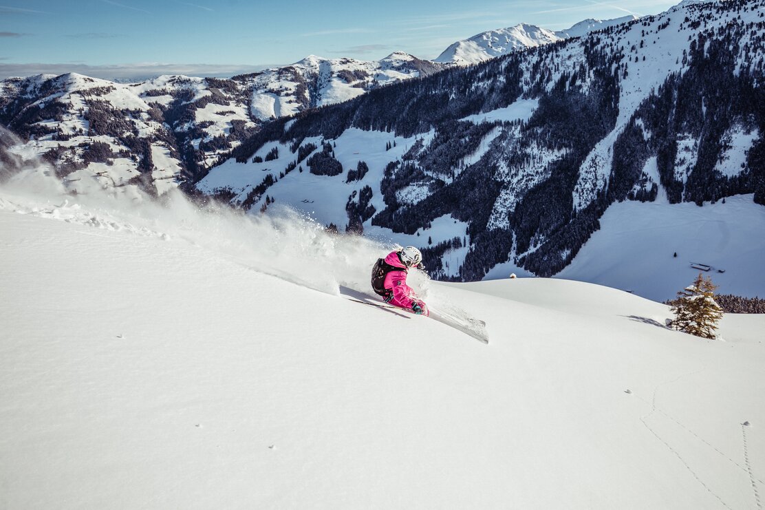 Eine Frau im pinken Skianzug fährt den Berg entlang, im Tiefschnee runter, im Hintergrund die Winterlandschaft 