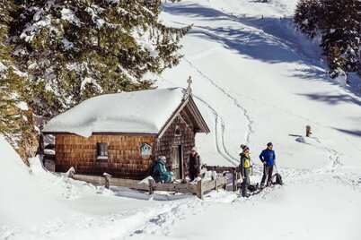 Eine Gruppe von vier Personen sitzt vor einer kleinen verschneiten Kapelle in Alpbach | © Alpbachtal Tourismus | shootandstyle 