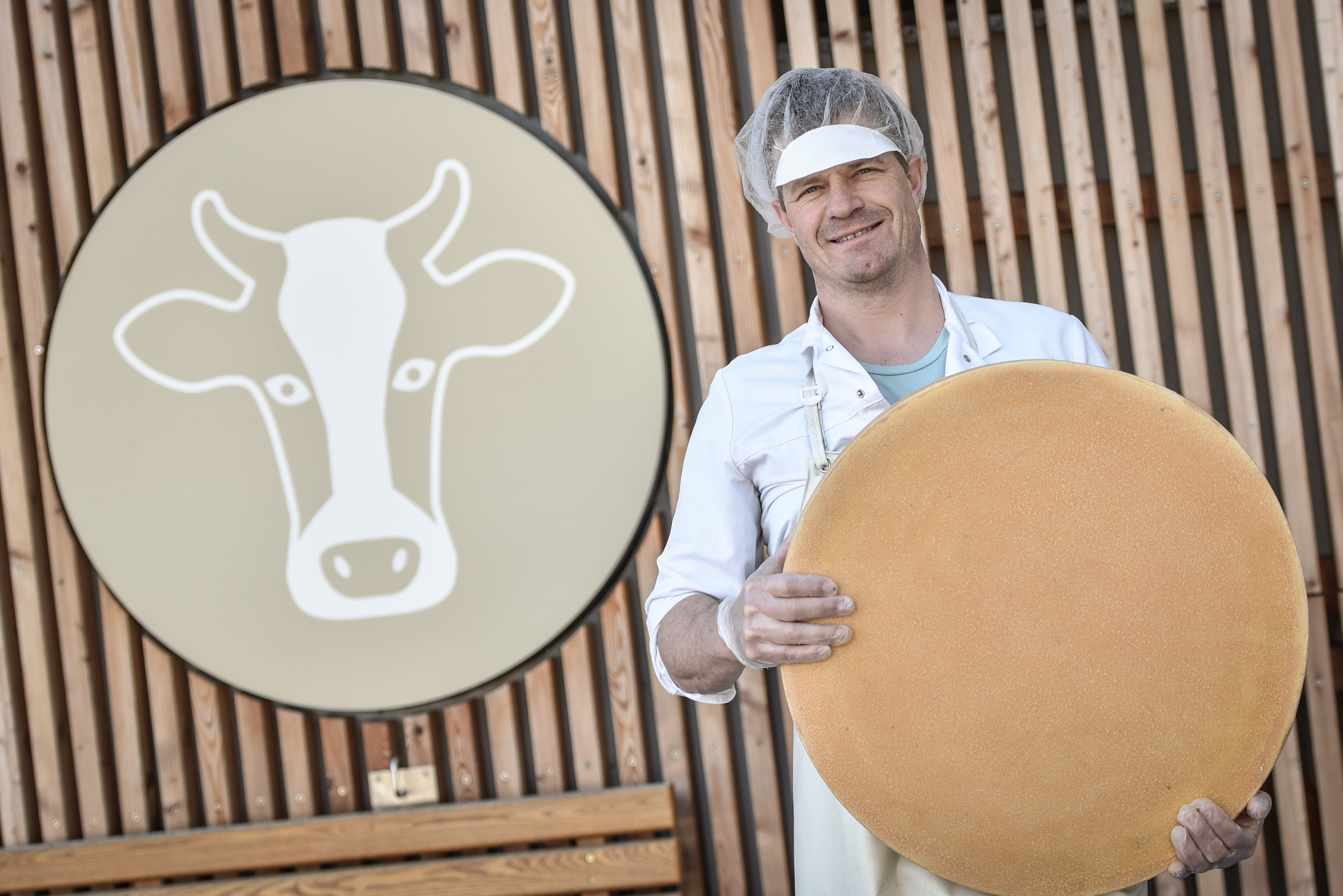 Ein Mann steht mit einem großem Leib Käse vor dem Logo der Alpbachtaler Heumilchkäserei in Reith i.A. | © Alpbachtal Tourismus