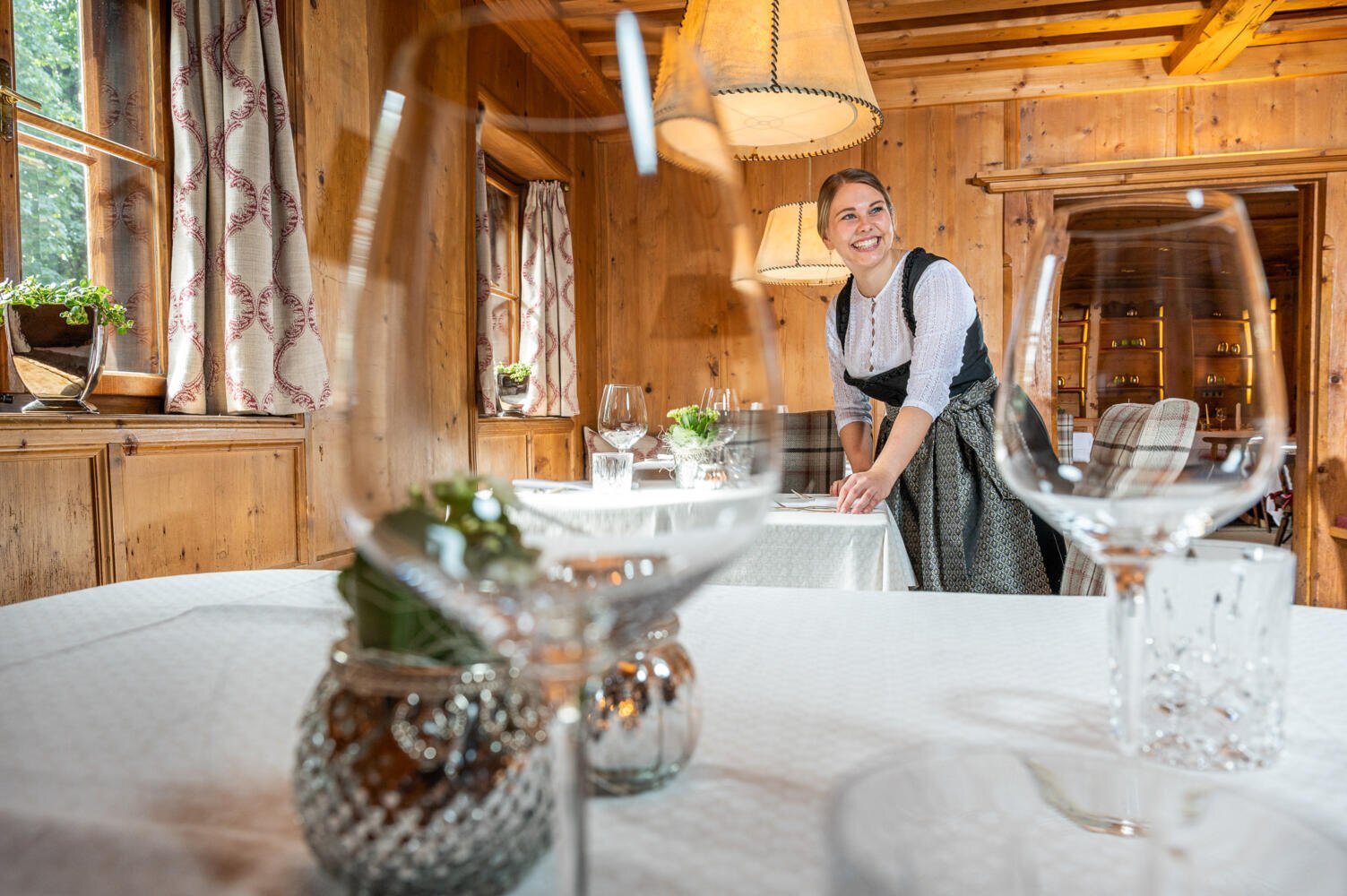 Im Vordergrund zu sehen sind Weingläser, im Hintergrund eine Kellnerin mit freundlichem Lächeln, beim Tisch eindecken | © Hotel Böglerhof 