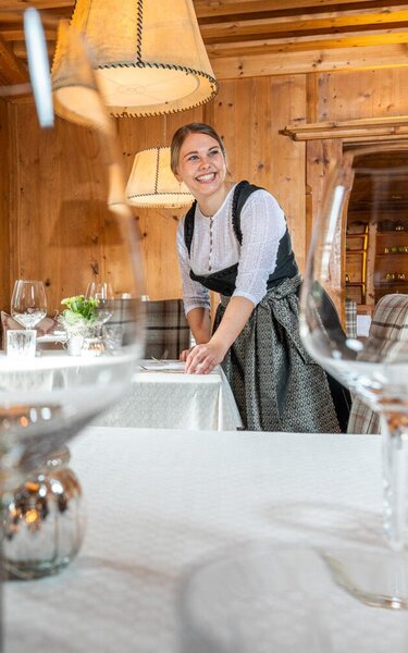 Im Vordergrund zu sehen sind Weingläser, im Hintergrund eine Kellnerin mit freundlichem Lächeln, beim Tisch eindecken | © Hotel Böglerhof 