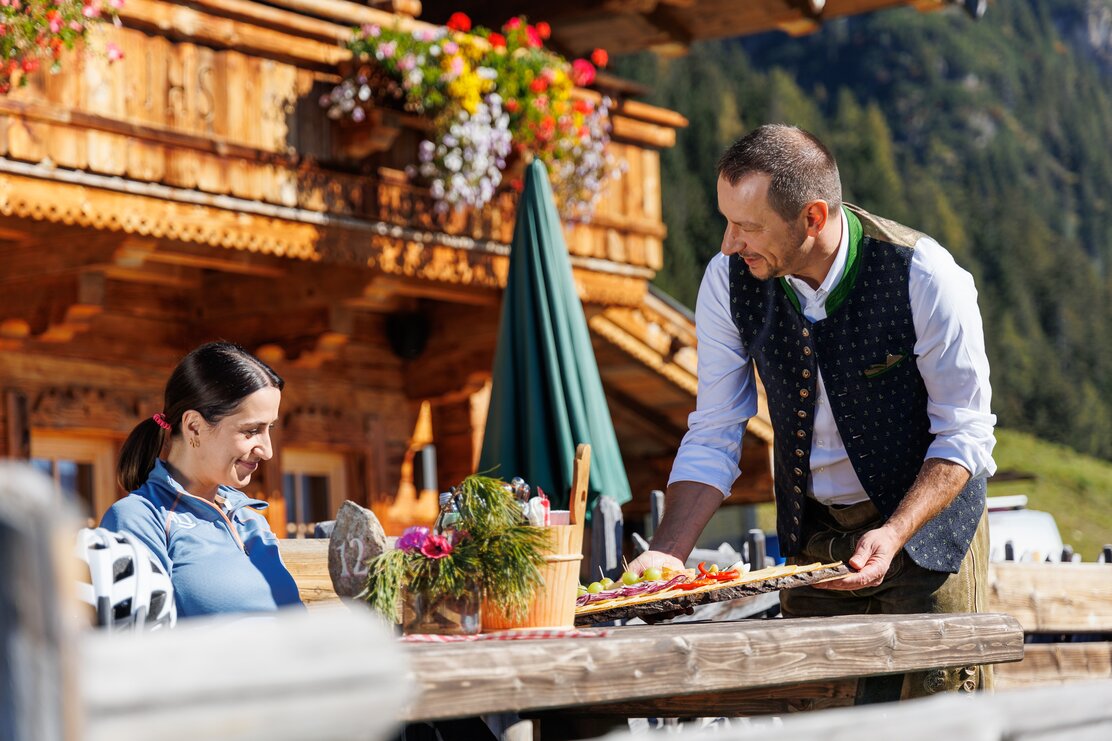 Ein Kellner bringt einer Frau eine Hüttenjause, auf der Bischofer Käsealm in Alpbach  | © Alpbachtal Tourismus | Maximalgrafik 