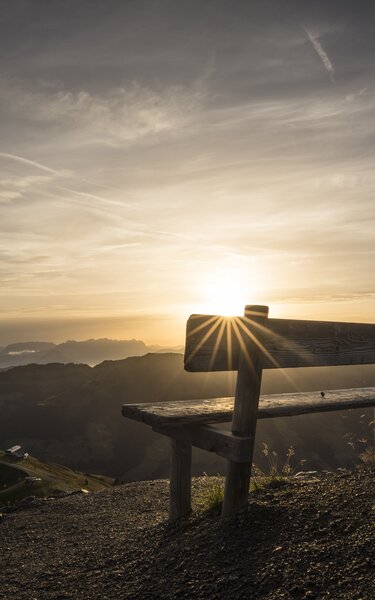 Eine Bank im Vordergrund, im Hintergrund Sonnenaufgang mit Blick auf die Wiedersbergerhornbahn | © Alpbachtal Tourismus | shootandstyle