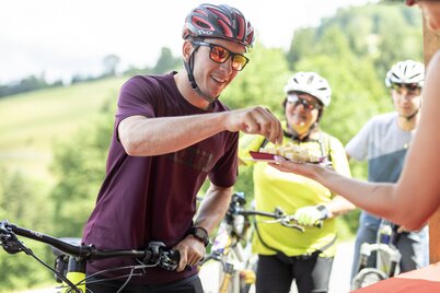 Ein Mann nimmt von einem Teller ein kleines Stück Käse herab, er hält nebenbei sein E-Bike fest  | © Alpbachtal Tourismus | shootandstyle 